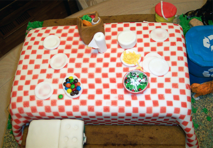 This is a closeup of the picnic table's edible fixns: grocery bag, plates and paper towels all on a fondant checkerboard-colored tablecloth. As the party was yet to arrive, the only other food on this table of chocolate cake is a plate of nachos, a plate of salad (for good nutrition) and some mini M&Ms — how appropriate for this sweet-tasting picnic-table cake. You'll note also the water cooler on the back gingerbread bench; the food chest is on the front bench. (7/2009)